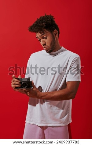 young african american man looking at his vintage camera on red background, photography as hobby