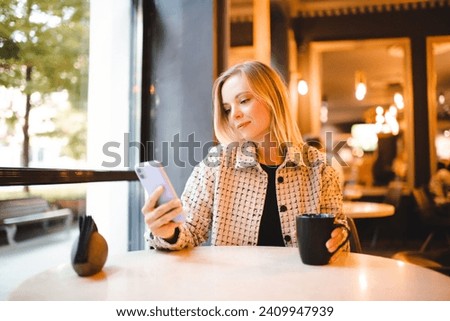 Beautiful blonde woman 24-25 year old drinking coffee in cafe over light looking in phone and typing messages