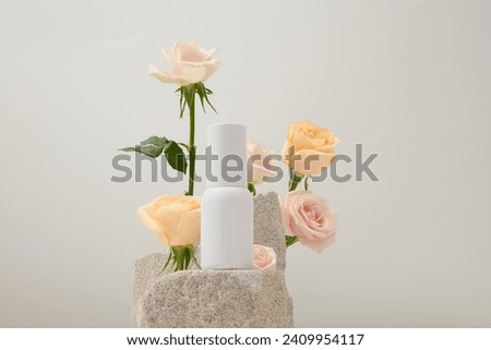Fresh roses are arranged around a gray stone slab with an unlabeled cosmetic bottle on a white background. Scene for cosmetics advertising. Front view. Royalty-Free Stock Photo #2409954117