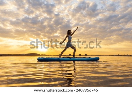 athletic woman doing YOGA on a inflatable SUP board on through shining water surface. aesthetically wide shot. Fitness at sunset on a lake
