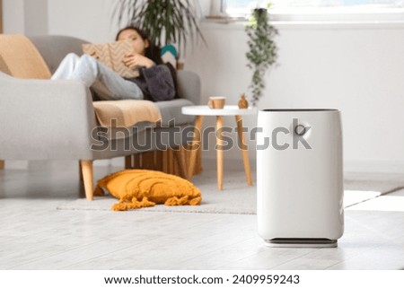 Air purifier against blurred woman reading book on sofa in living room Royalty-Free Stock Photo #2409959243
