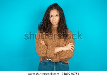 Gloomy dissatisfied Beautiful arab teen girl with curly hair wearing knitted sweater looks with miserable expression at camera from under forehead, makes unhappy grimace Royalty-Free Stock Photo #2409960751