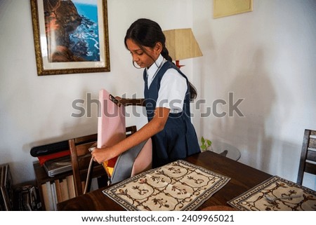 A pre-teen Indian girl wearing a blue and white school uniform packs her school bag for the day while standing at the dining table at her home in Mumbai, India.