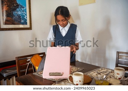 A pre-teen Indian girl wearing a blue and white school uniform packs her school bag for the day while standing at the dining table at her home in Mumbai, India.