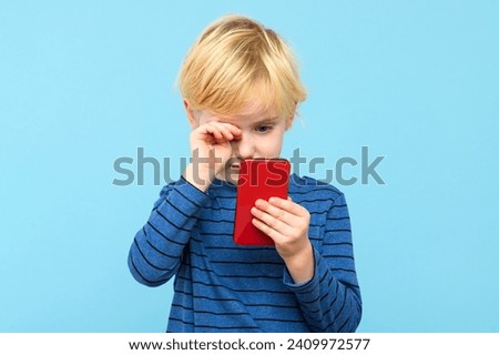 Young child holding smartphone, watching the screen, rubbing his eyes, isolated over pastel blue background. Children and technology, screen time control and eye health. Royalty-Free Stock Photo #2409972577
