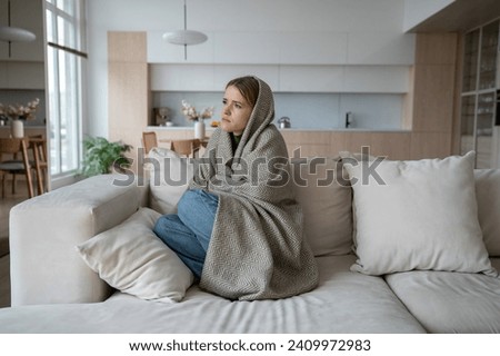 Sad tired woman sits on couch at home wrapped in plaid in living room. Blue mood of depressed girl boring. Nervous female feeling frustration hiding from society in loneliness solitude. Life problems. Royalty-Free Stock Photo #2409972983
