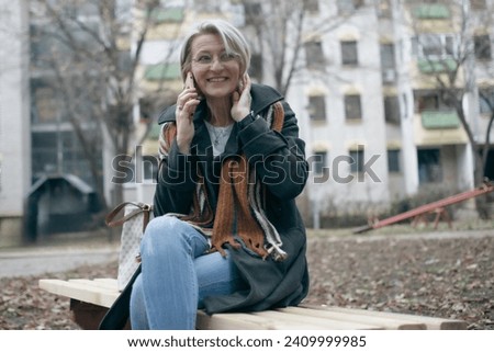 mature blond woman sitting on a park bench in front of a building on a cloudy winter day and talking on a mobile phone