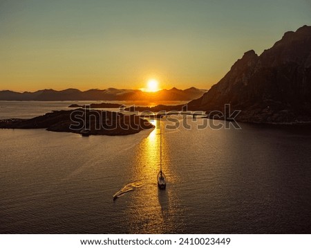 
A sailboat under the midnight sun, behind a large hill in Lofoten, near Henningsvaer, in the distance tall mountains, sun setting. Royalty-Free Stock Photo #2410023449