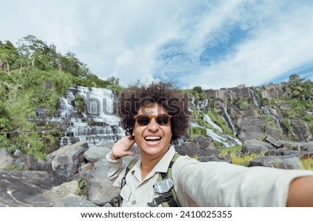 Young man excited hiker smiles while taking a selfie with waterfalls as background