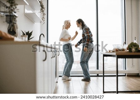 Elderly pensioners blond wife and husband with gray beard listening to music and dancing twist after delicious breakfast in bright modern spacious kitchen with panoramic windows.