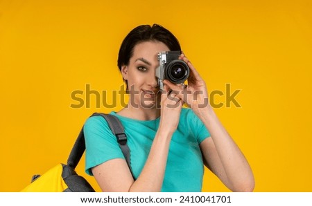 smiling woman photographer, a travel blogger with backpack taking photos with camera,  studio shot,  yellow background.