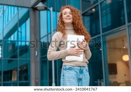 Portrait of beautiful happy young woman graduate with curly red hair holding laptop and excited  about news regarding her job application