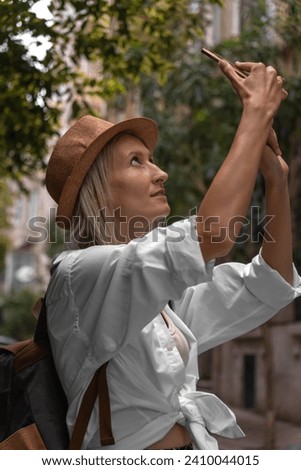 Stylish young blonde woman in a hat, white shirt with a backpack takes pictures on a beautiful street in the city on a mobile phone. Young female traveler. Vertical photo