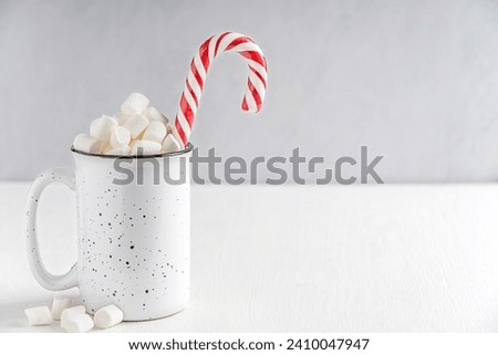 Homemade sweet traditional hot chocolate or cocoa drink decorated with marshmallow topping and striped candy cane served in ceramic mug or cup on white wooden table with copy space for cozy breakfast