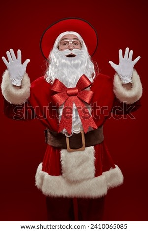 Fashion, Christmas and New Year. Modern Santa Claus in a traditional suit and a stylish wide-brimmed hat and a big red bow on his beard poses on a red studio background.  Royalty-Free Stock Photo #2410065085