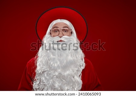 Modern handsome Santa Claus in a traditional suit and a stylish wide-brimmed hat on a red studio background. New Year's and Christmas. Christmas and fashion. Copy space.  Royalty-Free Stock Photo #2410065093