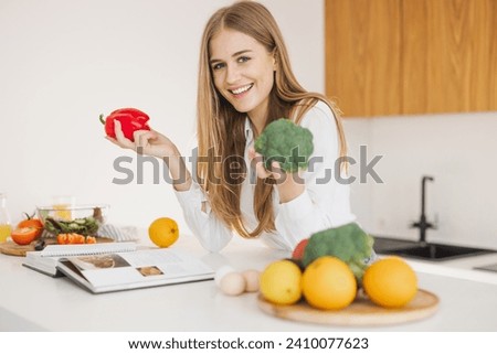 Portrait of a smiling cute blonde girl holding bell pepper and broccoli and reading recipe book in kitchen on table among cooking ingredients.