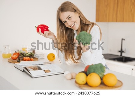 Portrait of a smiling cute blonde girl holding bell pepper and broccoli and reading recipe book in kitchen on table among cooking ingredients.