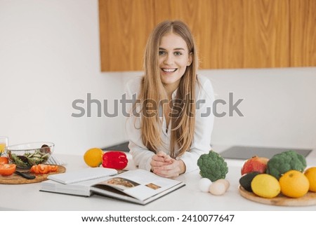 Portrait of a cute blonde girl smiling and reading a recipe book in the kitchen on the table among ingredients for cooking.
