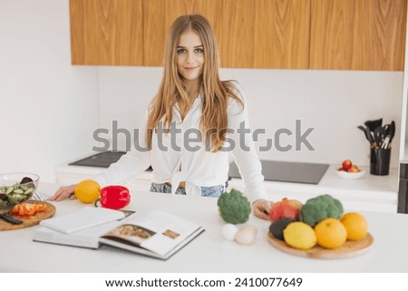 Portrait of a cute blonde girl smiling and reading a recipe book in the kitchen on the table among ingredients for cooking.