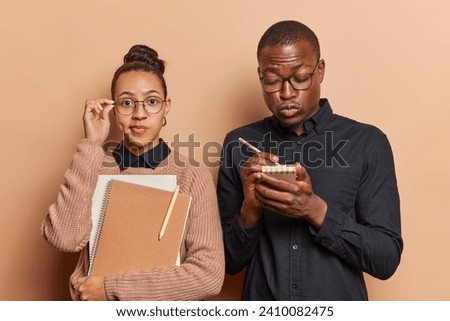 Woman and man students being busy preparing to exams write down notes in notepads dressed in casual clothes have serious expressions stand closely to each other isolated over brown background