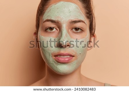 Self care and skin rejuvenation concept. Close up shot of serious woman applies green nourishing clay mask on face undergoes beauty treatments focused at camera poses against brown background.