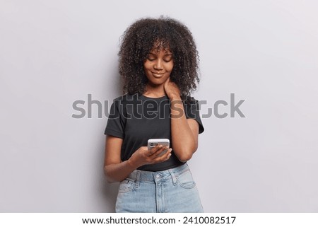Horizontal shot of serious curly haired young woman focused at smartphone screen browses internet searches information online wears casaul black t shirt and jeans isolated over white background.