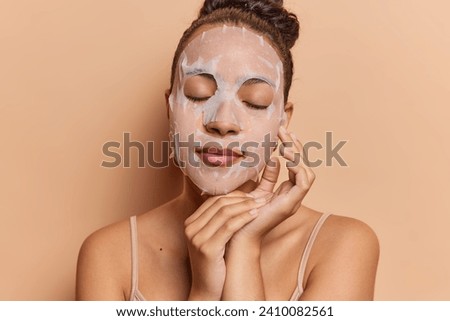 Beautiful dark haired woman applies beauty sheet mask on face for skin treatment touches face gently keeps eyes closed stands bare shouldered isolated over brown background. Wellness and wellbeing