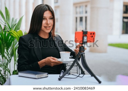 Engaged businesswoman conducting a live session, sipping coffee and interacting with an audience through a smartphone on a tripod