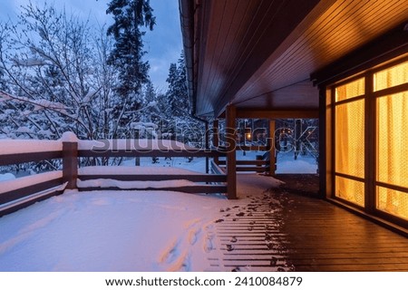 The atmosphere of peace. Light from the window of a wooden house on a snow-covered terrace. Twilight. Winter landscape. Royalty-Free Stock Photo #2410084879