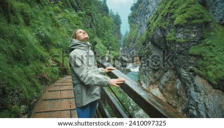 Woman enjoys the rain while hiking in a rocky gorge with mountain fast flowing river. Narrow wooden staircase along the canyon in Austria. Royalty-Free Stock Photo #2410097325