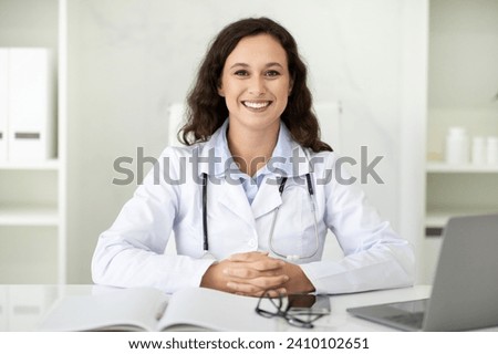 Portrait of cheerful beautiful millennial woman wearing medical coat doctor general practitioner sitting at desk in front of laptop computer in her office at clinic, smiling at camera Royalty-Free Stock Photo #2410102651