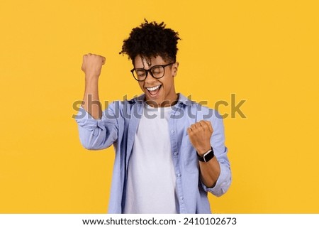 Ecstatic young black male student with glasses clenches fists in victory, eyes closed in elation, against vibrant yellow backdrop, symbolizing triumph Royalty-Free Stock Photo #2410102673