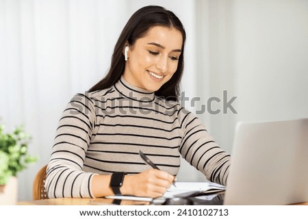 Focused young woman with wireless earbuds works diligently at her laptop, taking notes, exemplifying productive home office work in a bright setting. Study, online lesson, work