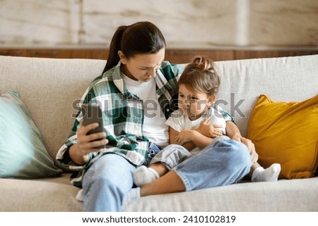 Mother showing to little daughter online content on smartphone at home, upset toddler girl frowning while watching cartoons with mom, sitting together on couch in living room interior, free space