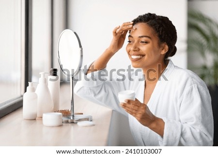 Beautiful Black Woman Sitting Near Mirror And Applying Moisturising Cream On Face, Attractive African American Lady Wearing Bathrobe Nourishing Skin After Bath, Making Beauty Routine At Home Royalty-Free Stock Photo #2410103007