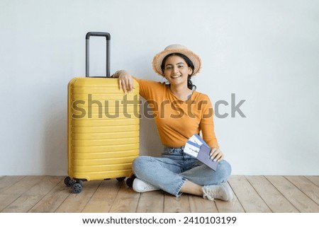 Smiling indian woman sitting on the floor beside her yellow suitcase, happy eastern lady wearing straw hat holding passport and tickets, expressing excitement of upcoming travels, copy space