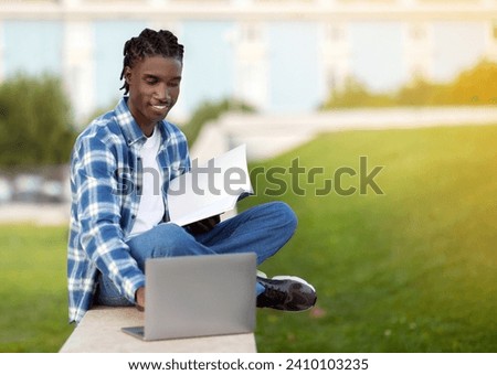 African American student guy websurfing on laptop holding workbooks and websurfing, studying sitting on grass outside university campus, immersed in digital learning leisure. Copy space Royalty-Free Stock Photo #2410103235