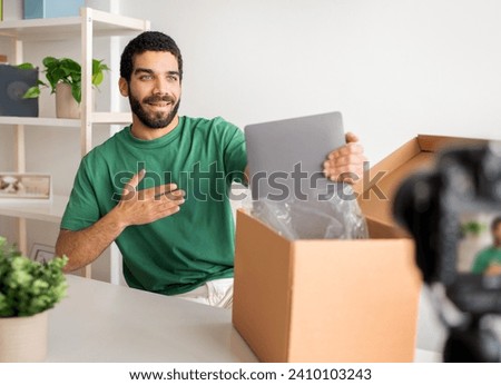 Long stock description: Smiling man in green shirt excitedly unboxing a new silver laptop at a tidy workspace with a camera filming, embodying a fresh start and modern technology