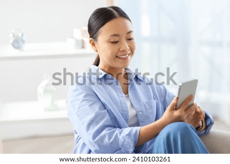 Positive attractive young chinese woman using phone at home, sitting on couch in cozy living room, reading blog online, websurfing, scrolling, chatting with men on dating mobile app, copy space Royalty-Free Stock Photo #2410103261