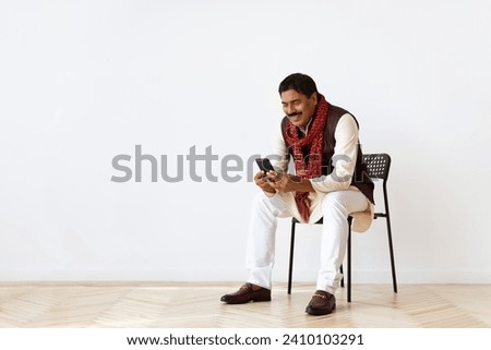 Handsome hindu man with moustache wearing indian clothing sitting on chair, using cell phone and smiling, websurfing, looking for job online, reading news, white wall background, copy space Royalty-Free Stock Photo #2410103291
