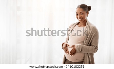 Positive expecting millennial african american woman hold hands in heart shape on her big tummy, smiling at camera. Happy young black pregnant lady posing next to window at home, banner, copy space Royalty-Free Stock Photo #2410103359