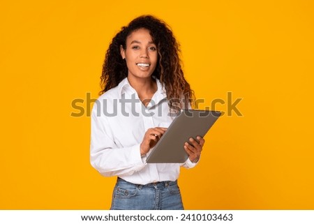 Cheerful African American lady engaging with digital tablet for freelance work in studio on yellow background. Lady in casual white shirt happily websurfing while posing with portable PC Royalty-Free Stock Photo #2410103463