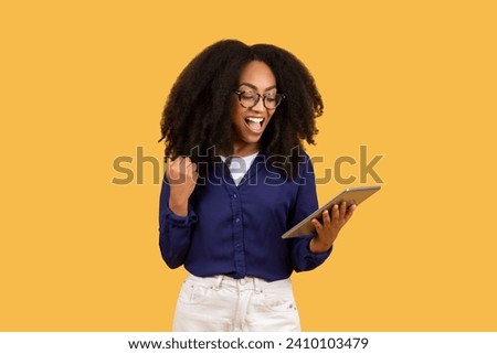 Elated young black woman with curly hair and glasses, holding tablet and making victory fist, exclaiming in triumph against yellow background