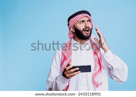 Upset muslim man wearing traditional islamic clothes loosing mobile game while enjoying online leisure in studio. Unhappy arab gamer in ghutra and thobe playing videogame on phone