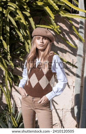 Woman model with tanned skin and blonde hair, in a vintage look wearing blue shirt, brown vest, hat and brown beige pants. Staying in front of a bunch of cactus. E-commerce, fashion photography  Royalty-Free Stock Photo #2410120505