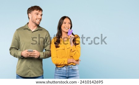 Smiling man holding smartphone and woman with credit card, possibly making an online transaction or planning a purchase, looking at free space on blue backdrop Royalty-Free Stock Photo #2410142695
