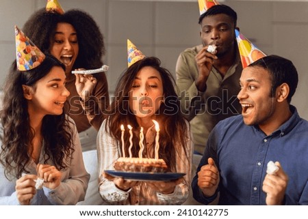Birthday party. Multiracial young friends group looking at bday woman blowing out candles on cake and making wish, their faces alight with happiness in festive indoor setting