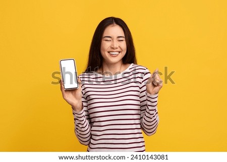 Overjoyed emotional young asian woman showing phone with white blank screen, grimacing and gesturing over yellow wall background, showing great online offer. Cashback