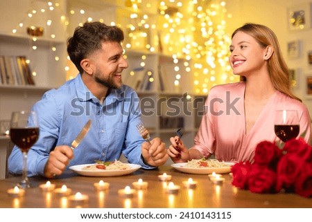 Cheerful couple sharing laugh over romantic dinner with red wine, pasta, and bouquet of roses, illuminated by warm candlelight, enjoying evening Royalty-Free Stock Photo #2410143115